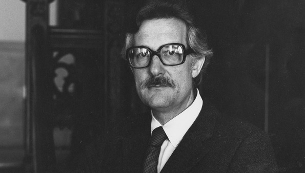 Hellmut Fritzsche, in suit, tie, and glasses, stands by a staircase.