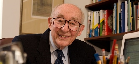Jack Halpern, wearing glasses, suit, and tie, smiles by a bookshelf.