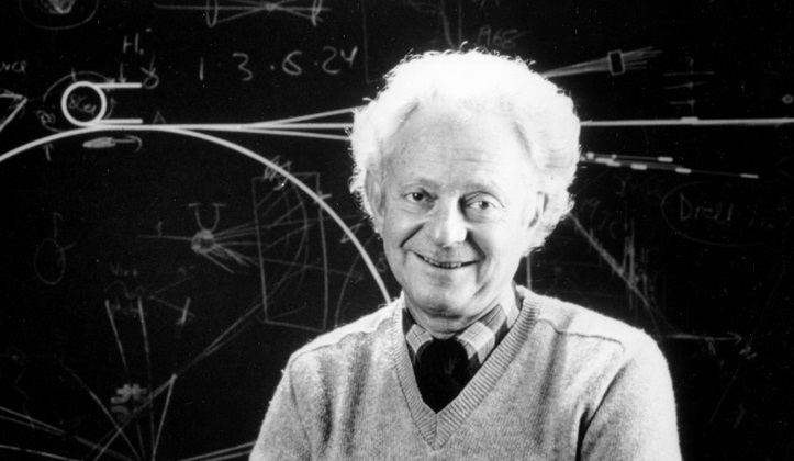 Leon Lederman, in tie and sweater, smiles in front of a chalkboard.
