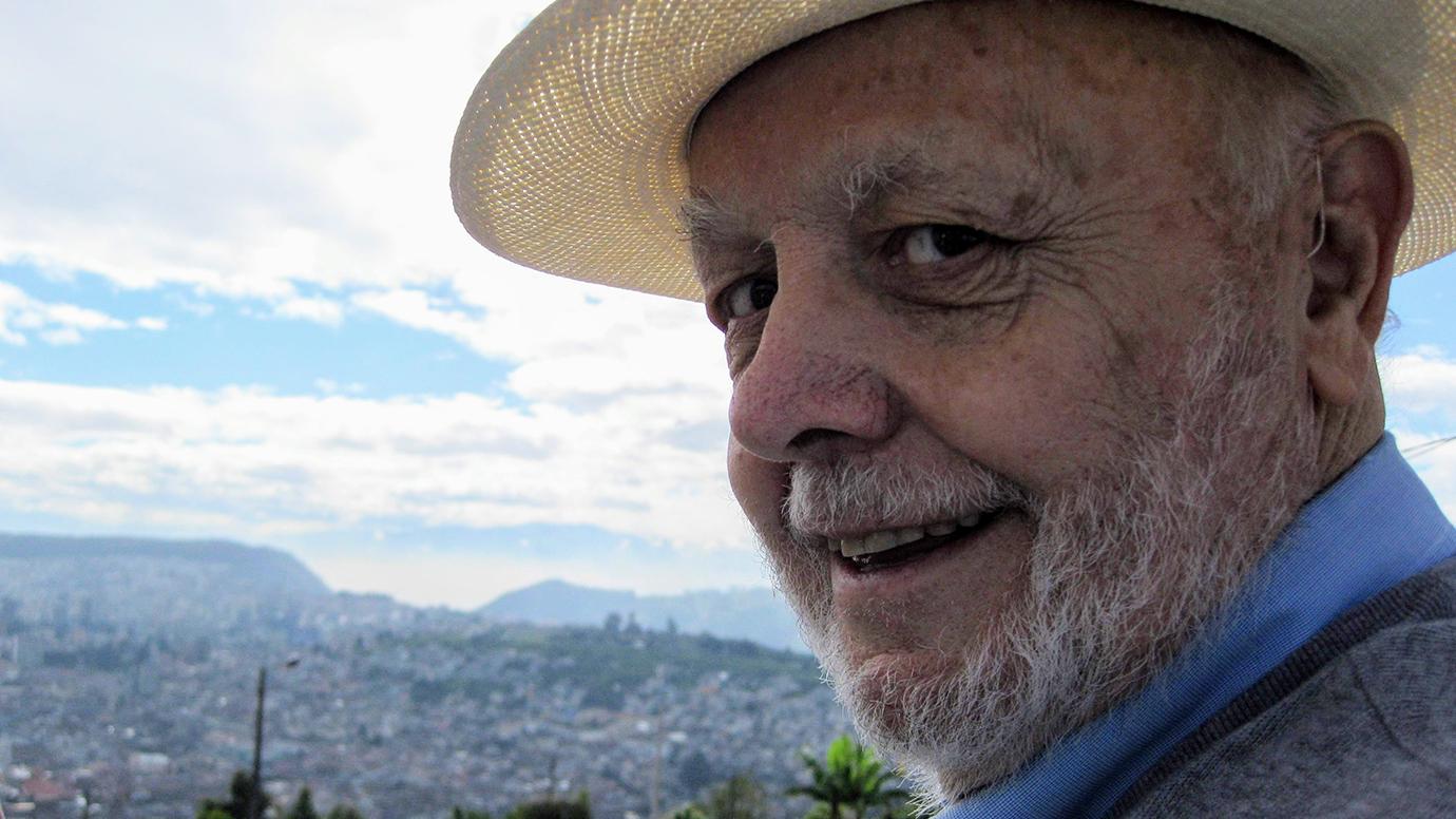 Max S. Bell, wearing a hat, turns to the camera as he stands before mountain scenery.