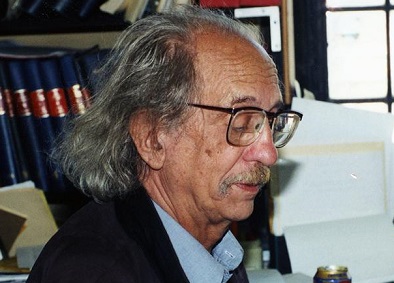 Miguel Civil, wearing glasses and a suit, sits by a bookshelf and window.