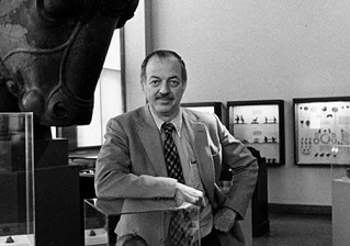 Robert McCormick Adams stands by a bull sculpture and glass boxes of artifacts.