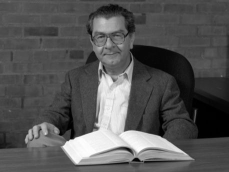 Victor Barcilon, facing camera, sits behind a desk with a book lying open on it.