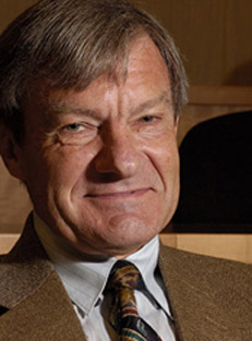 Abel Jeuland, wearing tan suit and dark tie, smiles as he stands in front of chalkboard. 