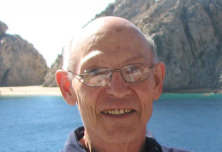 Selwyn W. Becker, wearing glasses, smiles while standing in front of ocean and mountain scenery.