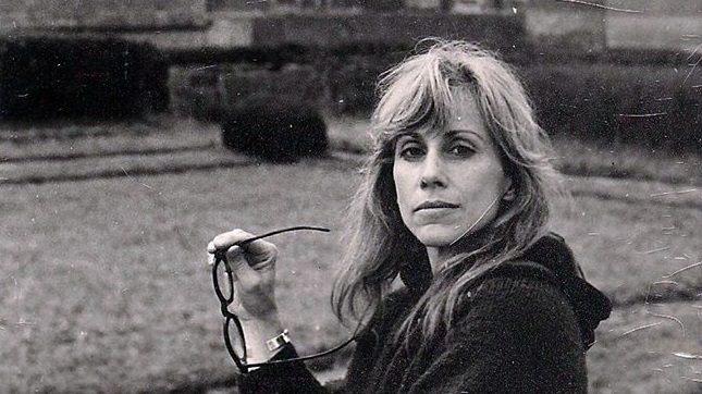 Anne Pippin Burnett, holding eyeglasses, sits on a bench near a field.