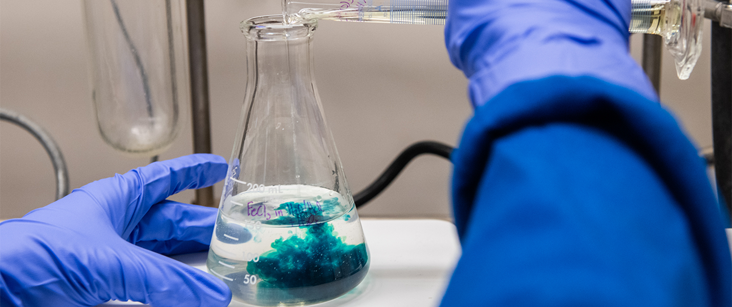 gloved hands pouring a chemical into another chemical, producing a blue color