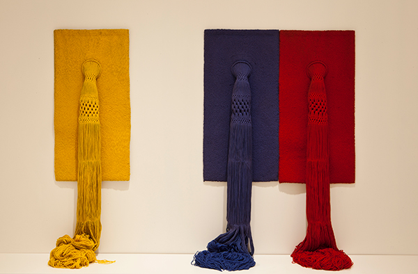 Yellow, blue, and red rectangles of yarn hang on a wall, with thick woven strands hanging down the center of each and gathering in a pool on the ground.