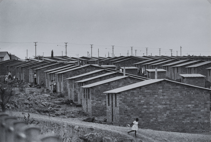 In a black and white photo, a girl in a white dress runs on gravel hill with rows of matching brick houses behind her.