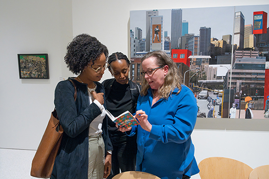Three people huddled around a book with artworks in a gallery behind them