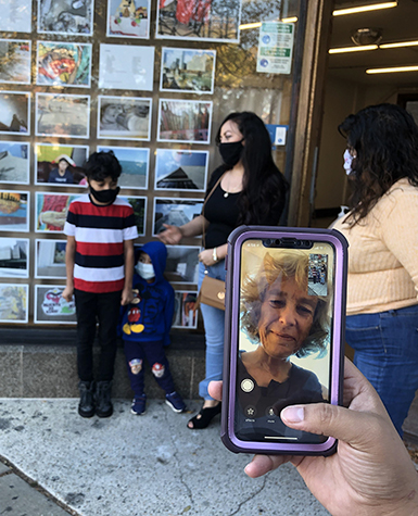 A family gathers in front of a storefront window full of photographs. In the foreground, a hand holds a phone up to share with a woman over video chat.