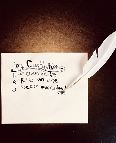 A quill pen rests next to a a piece of paper with calligraphy writing that reads: My Constitution, 1) Ice cream all day, 2) Kids can vote, 3) Soccer every day!