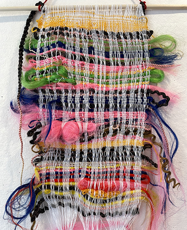 A roughly rectangular shaped weaving made of colorful yarn and braided thread hanging on a wooden dowel.